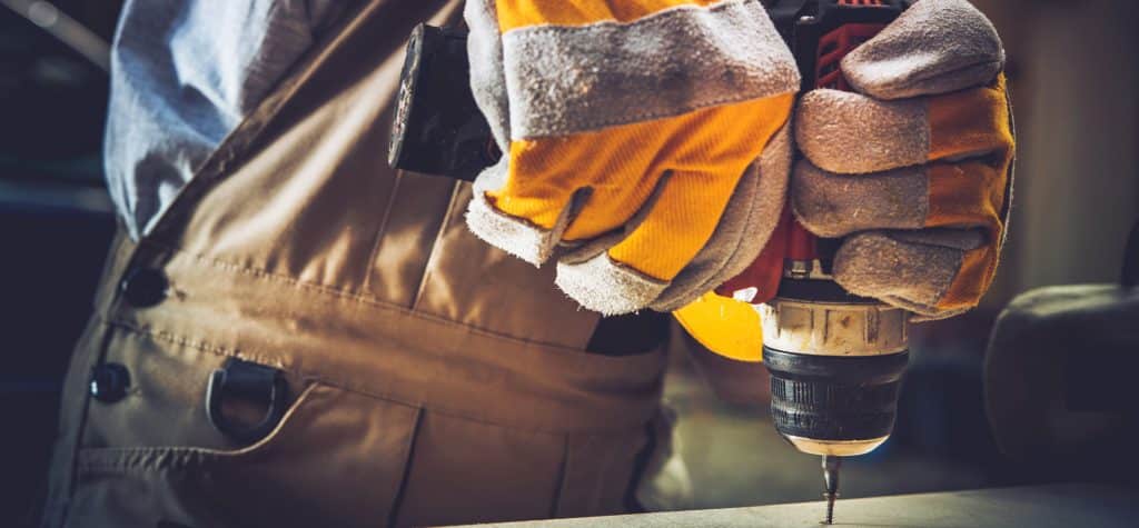 A person wearing heavy-duty gloves and overalls uses a power drill to screw into a wooden surface, with the focus on their hands and the drill—ideal for tasks like industrial gearbox rebuild or emergency gearbox repair.
