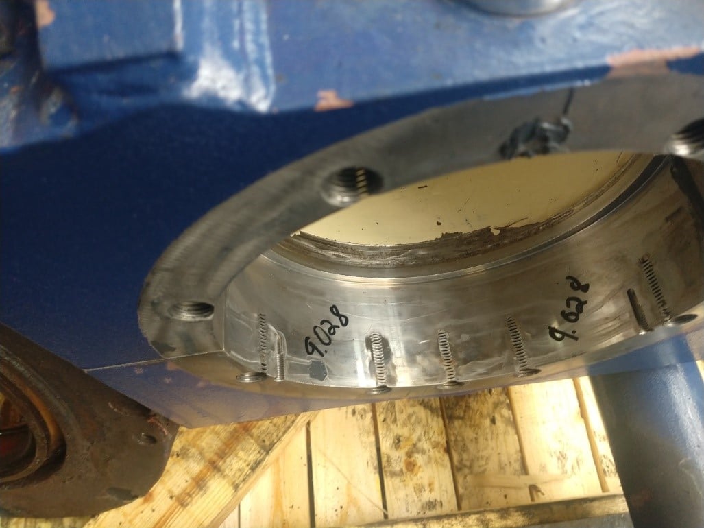 A close-up view of a metallic cylindrical machine part with visible bolts, handwritten markings, and slight wear, set against a blue housing and wooden surface—ideal for industrial gearbox repair or emergency gearbox repair.