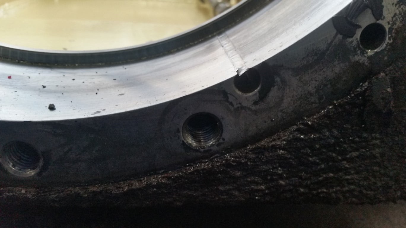 Close-up of a metallic circular surface from an industrial gearbox repair, featuring several threaded holes and visible scratches. The lower area is rough and dark, while the upper surface is smooth and shiny, showing signs of wear and residue.