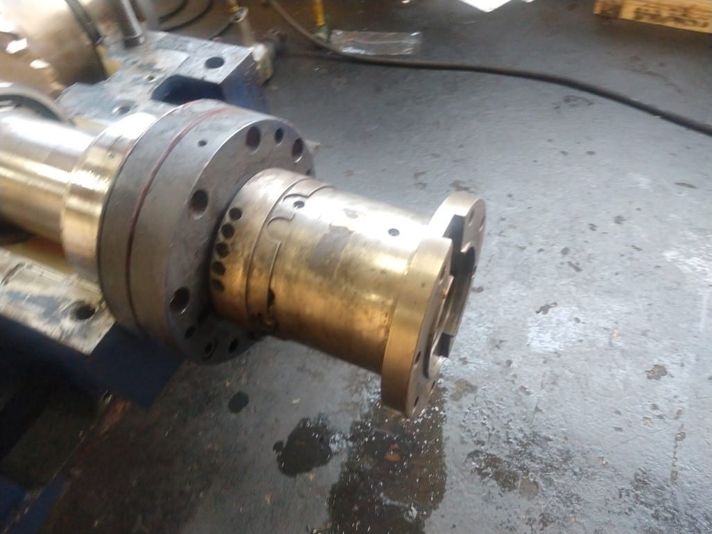 A close-up view of a large industrial metal shaft with coupling attached, placed on a workshop floor with some liquid spillage nearby. The background includes tools and equipment used in industrial gearbox repair.