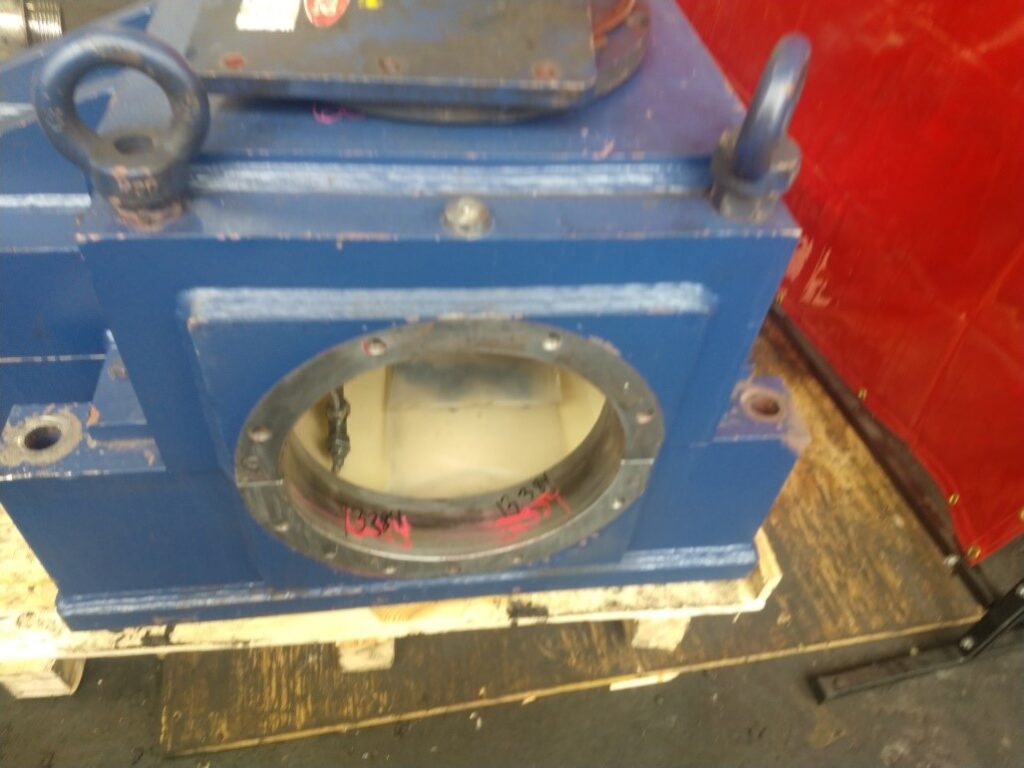 A large blue metal industrial component with a circular opening on one side, sitting on a wooden pallet. Ideal for those seeking industrial gearbox repair or rebuild services. The background features a red barrier and dark floor.