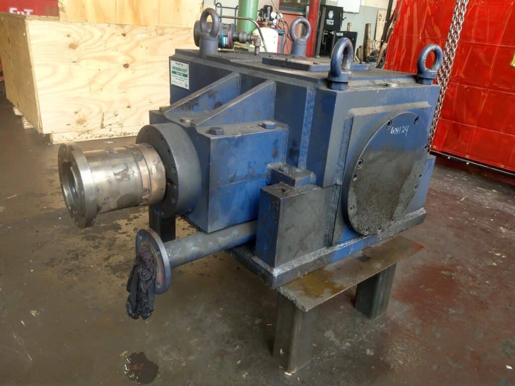 A large industrial blue gearbox sits on a workshop floor, showing signs of use with oil stains and grime. The cylindrical metal shaft and lifting eyes suggest it may soon undergo industrial gearbox repair or rebuild services.