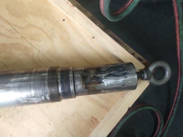 A close-up of a metallic cylindrical object with an eye bolt on one end, lying on a wooden surface. Nearby hoses and dark stains suggest recent emergency gearbox repair or industrial gearbox rebuild work.