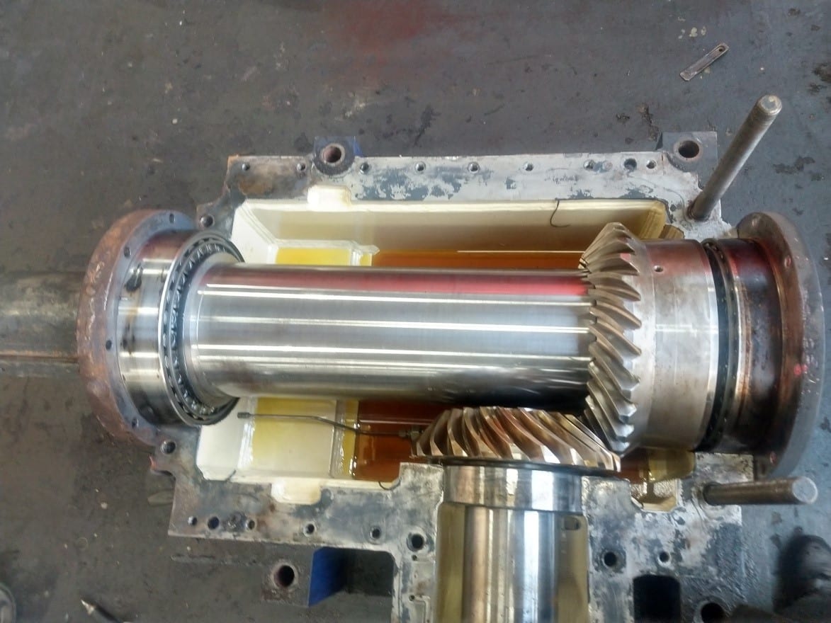 A close-up view of an open gearbox shows two interlocking metal gears and shafts with oil at the bottom of the housing, highlighting the precision involved in industrial gearbox repair within a workshop setting.