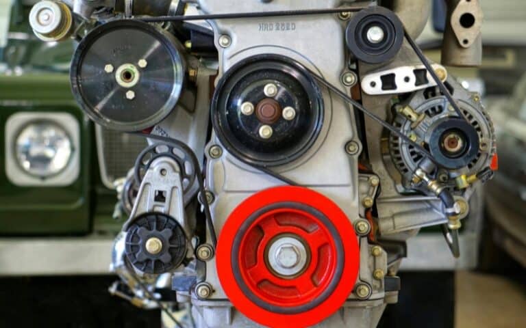Close-up view of a car engine with a prominent red pulley in the center, various belts and mechanical components visible—ideal for showcasing industrial gearbox repair—with a blurred green vehicle in the background.