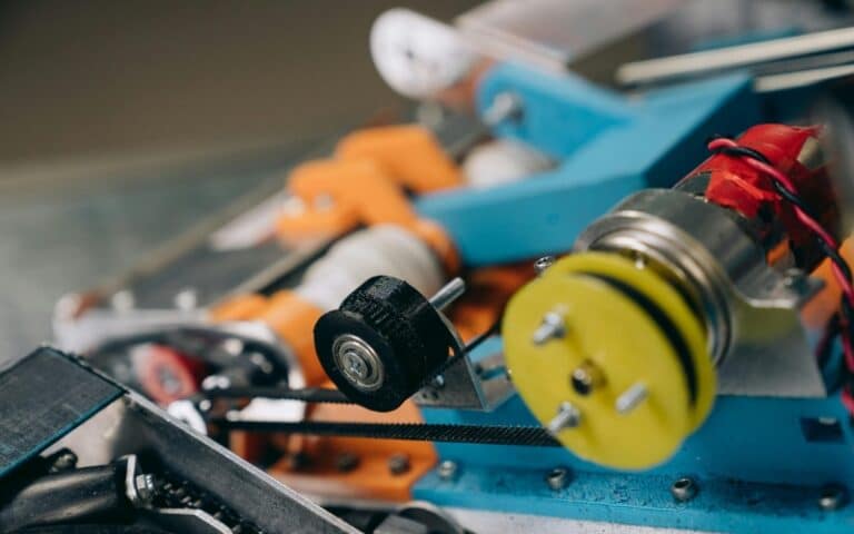 Close-up of a colorful robotic mechanism, featuring gears, pulleys, and wires—ideal for showcasing industrial gearbox repair or gearbox rebuild services. The components include metal, plastic, rubber parts with visible bolts and a black wheel on a belt system.