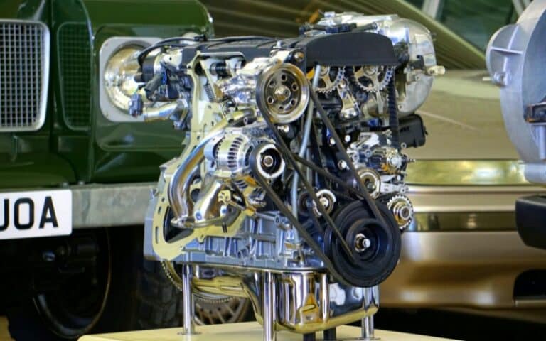 A detailed car engine model is displayed on a stand, showing visible belts, gears, and metallic components—ideal for illustrating industrial gearbox repair—with classic cars blurred in the background.