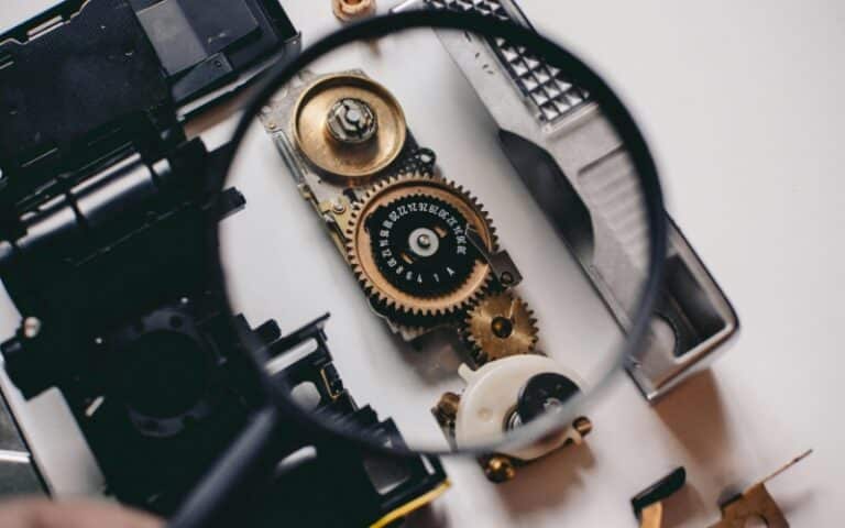 A close-up view of mechanical gears and parts, including a large brass gear and smaller components, seen through a magnifying glass on a white surface. The image suggests precision work like industrial gearbox repair or rebuild services.