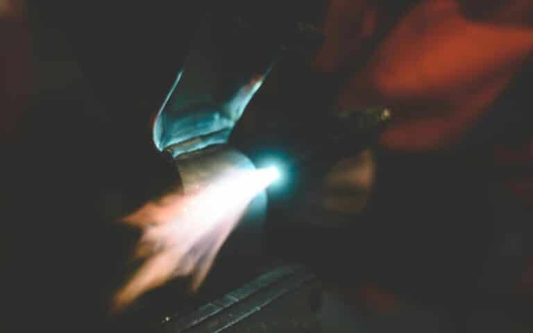 A close-up of a welding torch emitting a bright flame onto a metal surface, set against a blurred dark and orange background—capturing the intensity of emergency gearbox repair in action.