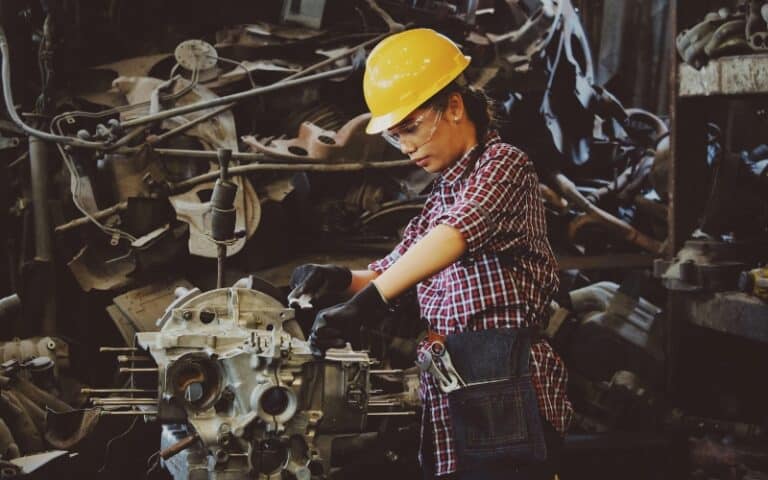 A woman wearing a yellow hard hat, safety glasses, and gloves works on an industrial gearbox repair in a workshop, surrounded by metal tools and engine components.