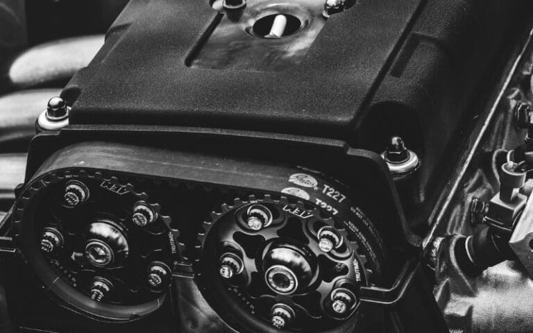 Close-up, black-and-white image of a car engine, focusing on the cam gears and timing belt, with bolts and mechanical components visible—highlighting the intricate details often addressed in industrial gearbox repair.
